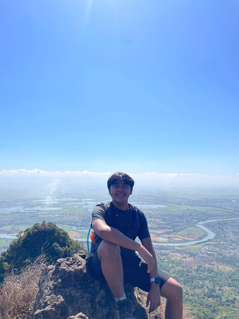 Bernard sitting on a rocky mountaintop overlooking a wide Philippine landscape.
